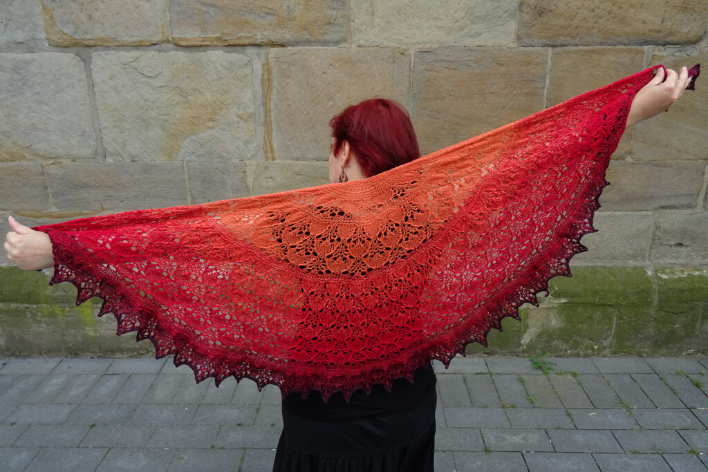 A half-circle shawl in a gradient from orange to dark red is spread out behind a white woman to show off the full shawl. The shawl features a dragon lace pattern and garter sections.