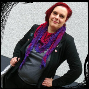 Photo is taking at an angle. It shows a white woman with bright red hair wearing grey and black clothes and a bright red and purple crochet scarf. She smiles lightly at the camera.