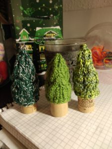Three knit pine trees with cable pattern on cork stems, each a different shade of green. In the background are Christmas decorations