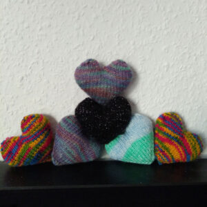6 handknit hearts in various colours are arranged in front of a white wall