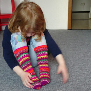 a red-haired child sits on the floor with knees drawn up, looking down and touching a pair of colourful, knee-high socks.