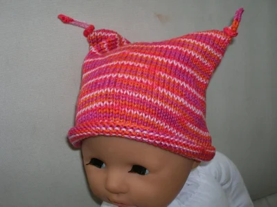 A baby doll wears a pink striped hat with a rolled brim and tied i-cords at the two points at the top