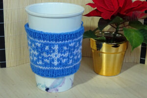 Free pattern - white cup with a blue coffee sleeve with white snowflake stands next to a small plant.