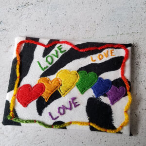 ATC with a rainbow of heart on zebra striped fabric. A thick strand of rainbow yarn frames the piece and the word LOVE is written in three places